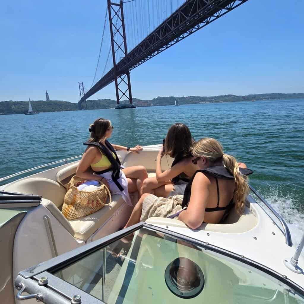 Grupo de amigas a bordo da lancha, navegando sob a Ponte 25 de Abril com vista para o Cristo Rei.